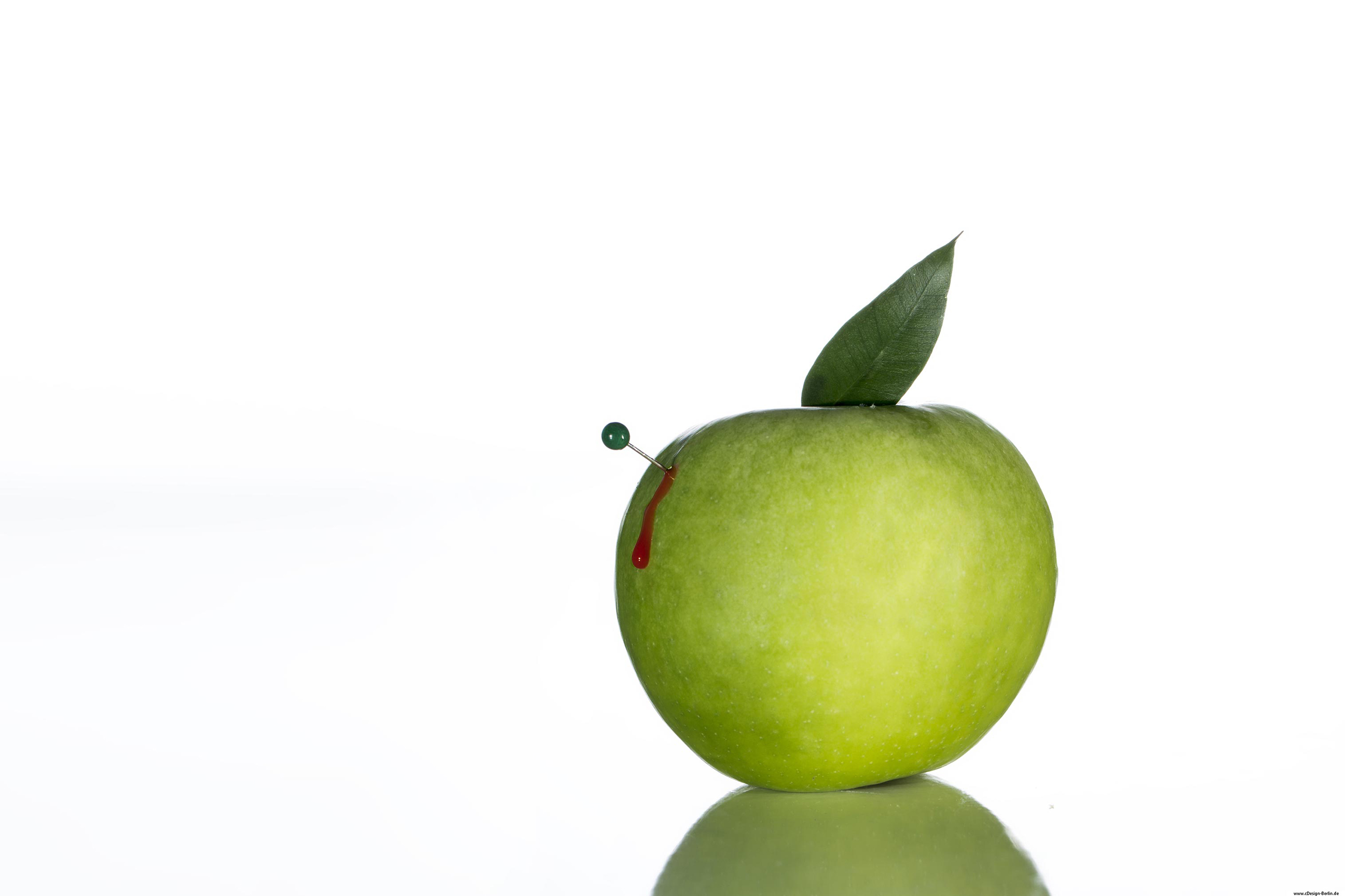Der Hintergrund auf diesem Bild ist weiß gehalten. Nicht  mittig, sondern im goldenen Schnitt ist ein grüner Apfel abgebildet, dieser spiegelt sich auf einer Glasplatte wider. Links im Apfel steckt eine Stecknadel. Der Apfel blutet, es soll die Verletzbarkeit der Natur symbolisieren.
