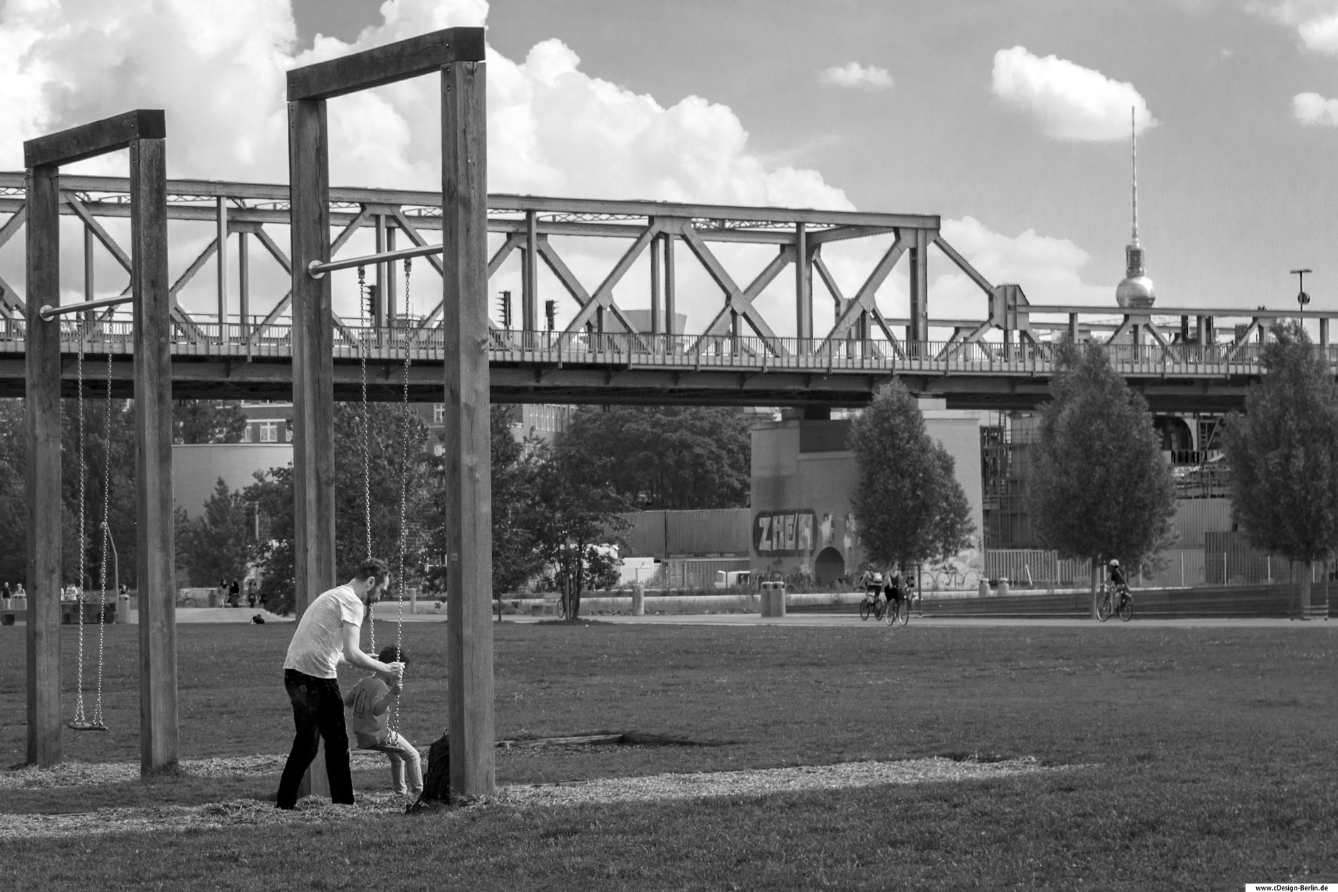 Komm Papa wir gehen Schaukeln. Dieses Foto entstand in einem belebten Park im Herzen Berlins. Am linken Bildrand sieht man einen Vater auf einer Wiese, der sein Kind beim Schaukeln behilflich ist. Seine Hände umklammern die Schaukel, sein Blick auf den Kopf seines Kindes gerichtet. Die Schaukel ist an zwei hohen Balken befestigt, rechts von ihnen befindet sich ebenfalls eine Schaukel, leerstehend.
In der Bildmitte ist eine Brücke zu sehen, dort führt die Berliner S-Bahn lang. Hinter der Brücke rechts vom Bild sieht man in einer Silhouette den Berliner Fernsehturm. Unter der Brücke ist ein Rad-Gehweg, darauf sind Fahrradfahrer zu sehen. Am oberen Bildrand sind der Himmel mit dicken Wolken zu sehen. Dieses Foto habe ich ebenfalls Schwarz/ weiß gehalten.
